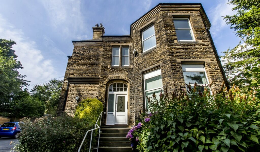 A stone two storey house with stone steps leading up to it, with railings beside them. Outside the house are hedges and trees.