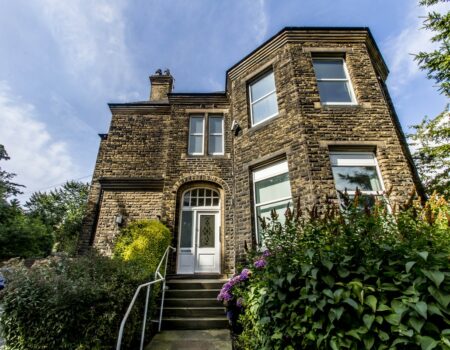 A stone two storey house with stone steps leading up to it, with railings beside them. Outside the house are hedges and trees.