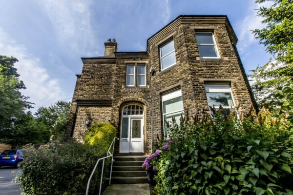 A stone two storey house with stone steps leading up to it, with railings beside them. Outside the house are hedges and trees.