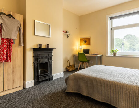 A bedroom with a grey carpet, light coloured wooden wardrobe, with a black fireplace in the center of the room. The bed is a double bed with grey and white bedding. There is a desk in the corner with a lit desk lamp, just to the left of a large square window facing the bed. The room is well decorated with frames on the wall and small house plants on the window sill.