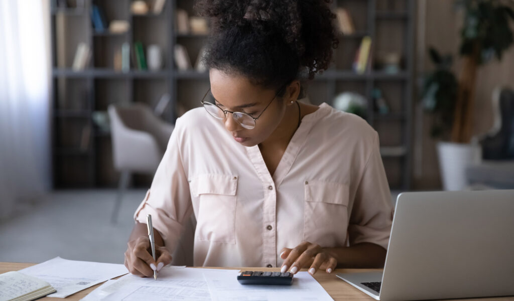 A mixed race person wearing glasses at a wooden desk writing with a pen on some paper, whilst using a calculator. There is a laptop to the right of them.